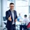 Portrait of businessman welcoming new employee to his business team and company, giving hand forward in modern office with colleagues in the background.