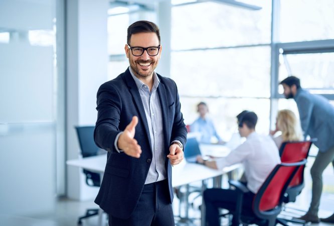 Portrait of businessman welcoming new employee to his business team and company, giving hand forward in modern office with colleagues in the background.